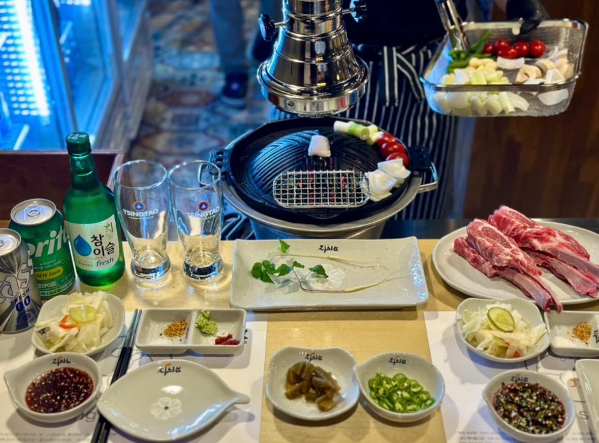 경상남도 창원시 징스램 상남점 다찌석 조명 아래 선홍빛 양갈비와 채소가 차려진 1인 화로구이. 소주와 음료가 나란히 놓인 혼술 식탁.