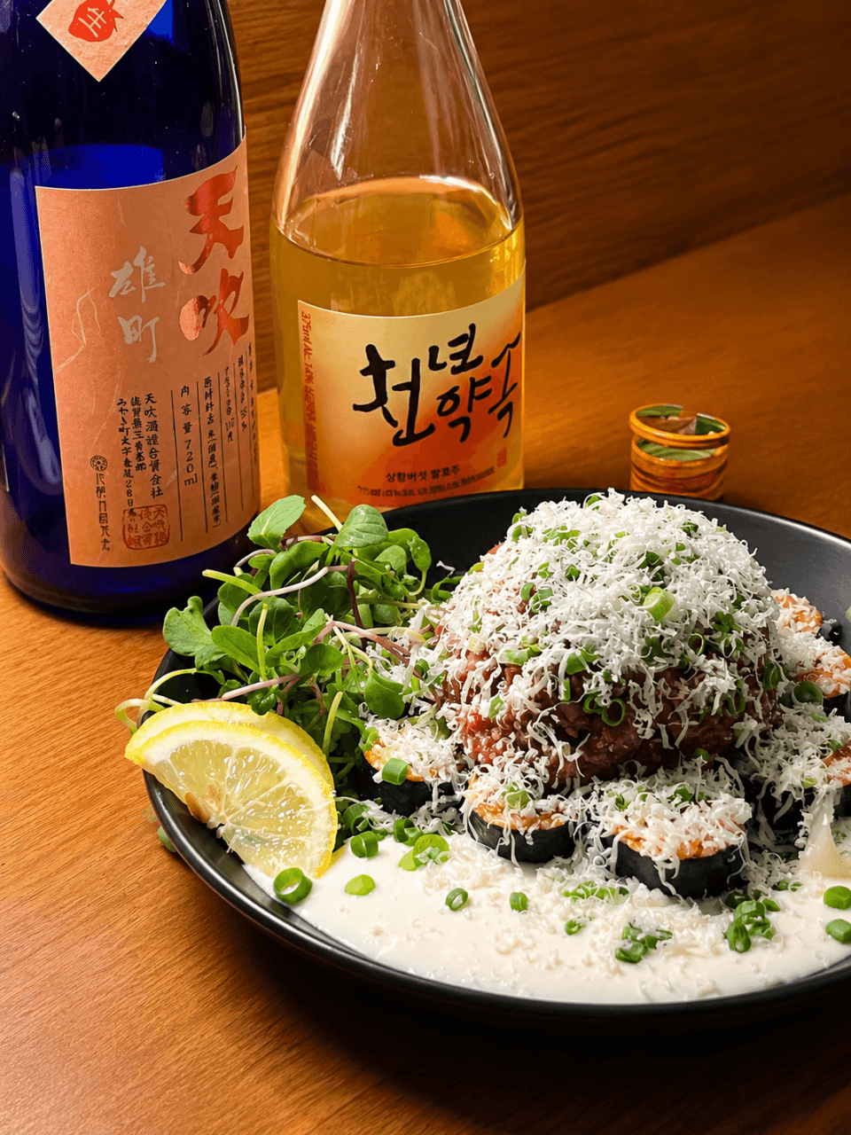 경상남도 창원시 순조실비 다찌석에 차려진 혼술 메뉴. 치즈가 내려앉은 육회와 김밥, 두 종류의 전통주 병이 조명 아래 나무 테이블 질감과 대비됨.