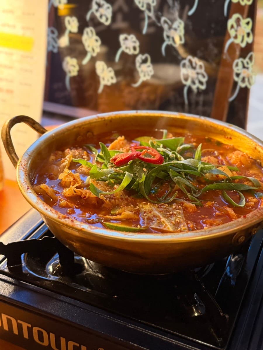 경상남도 창원시 순조실비 다찌석 1인 혼술 상황. 휴대용 버너 위 양은냄비에 담긴 붉은 찌개와 파채. 창가 조명 아래 놓인 1인 좌석과 기타 요리.