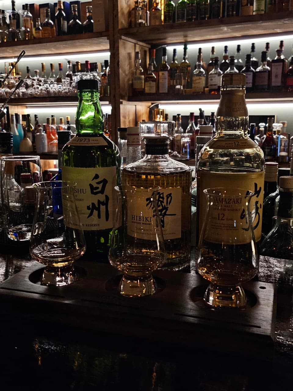 경상남도 진주시 미드나잇덴 바(Bar) 혼술 - 조명 아래 다찌석에 차려진 위스키 시음 세트와 테이스팅 글라스. 하쿠슈와 야마자키 병이 놓인 원목 카운터의 금빛 위스키 질감.