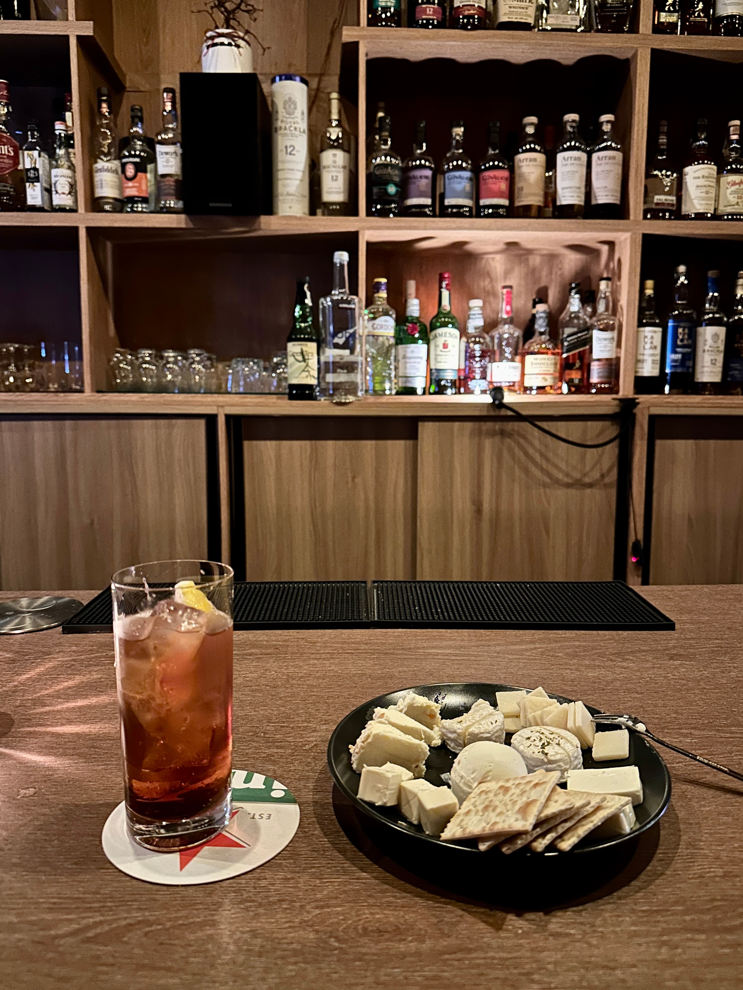 서울특별시 동작구 혼술 바(Bar) 귀로의 나무 다찌석 위로 붉은 칵테일과 치즈 크래커 플레이트가 놓여 있고 배경의 수많은 위스키 병들이 조명을 받는 모습