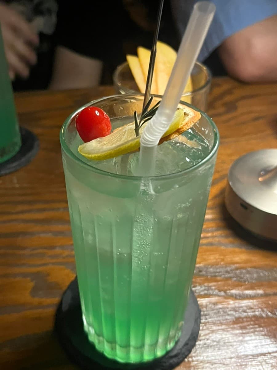 충청북도 청주시 제주아홉 혼술바 청주 지웰시티점 바(Bar) 다찌석 위 레몬과 체리가 올라간 초록색 하이볼. 낮은 조도의 나무 테이블에 놓인 1인용 음료와 안주 상황.