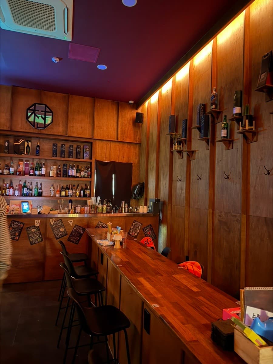 충청북도 청주시 청주 혼술바 해방촌 바(Bar)에서 오렌지빛 간접 조명이 비추는 목재 다찌석에 앉아 혼술을 즐기는 상황. 벽면 선반에 진열된 위스키병과 1인 좌석이 배치됨.