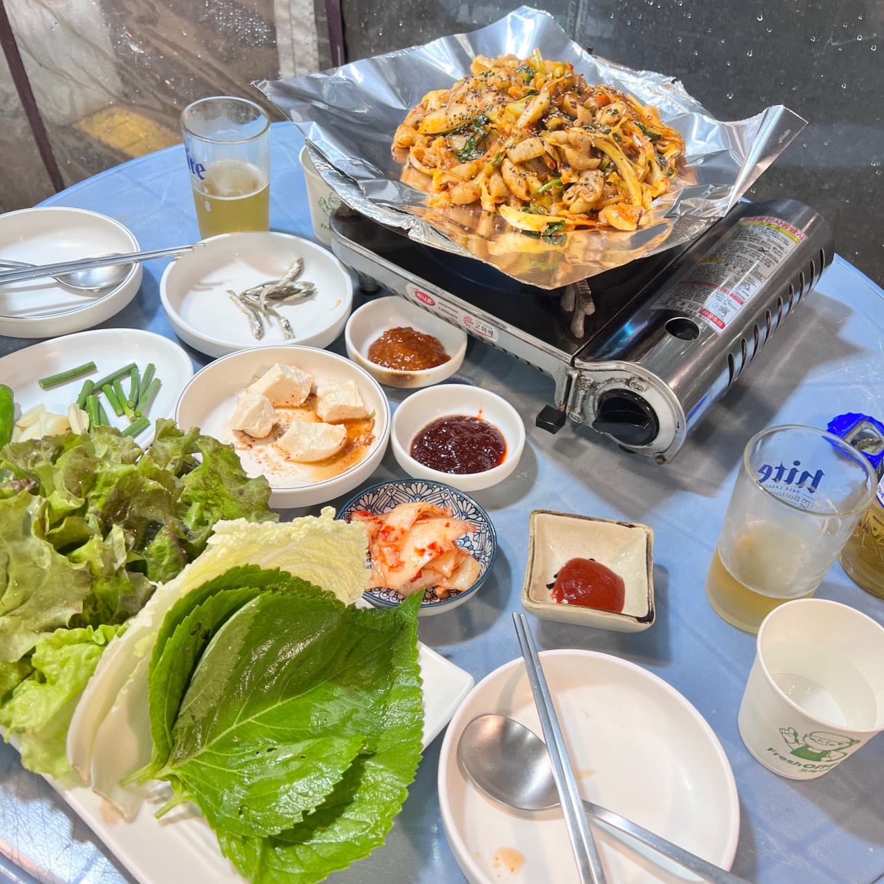 서울특별시 동작구 맥주 호프 그냥갈수없잖아 - 파란 원형 테이블 위 휴대용 가스레인지에 차려진 곱창볶음과 신선한 쌈 채소로 구성된 1인 혼술 상차림.