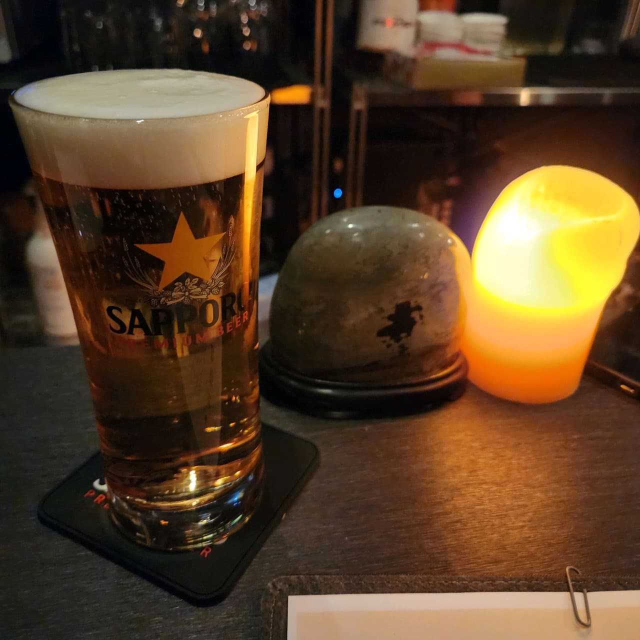 서울특별시 동작구 혼술 바(Bar) 사무실 - 다찌석에 놓인 거품 가득한 삿포로 생맥주와 캔들 조명. 어두운 배경의 1인 테이블 구성.