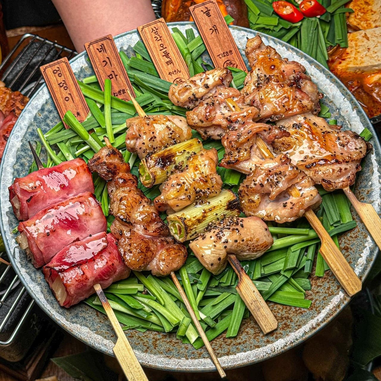 충청북도 청주시 숯토리 청주강서점 이자카야 다찌석의 1인 꼬치 세트. 부추 위에 놓인 윤기 나는 야키토리와 베이컨 말이, 나무 이름표가 차려진 혼술 상차림.